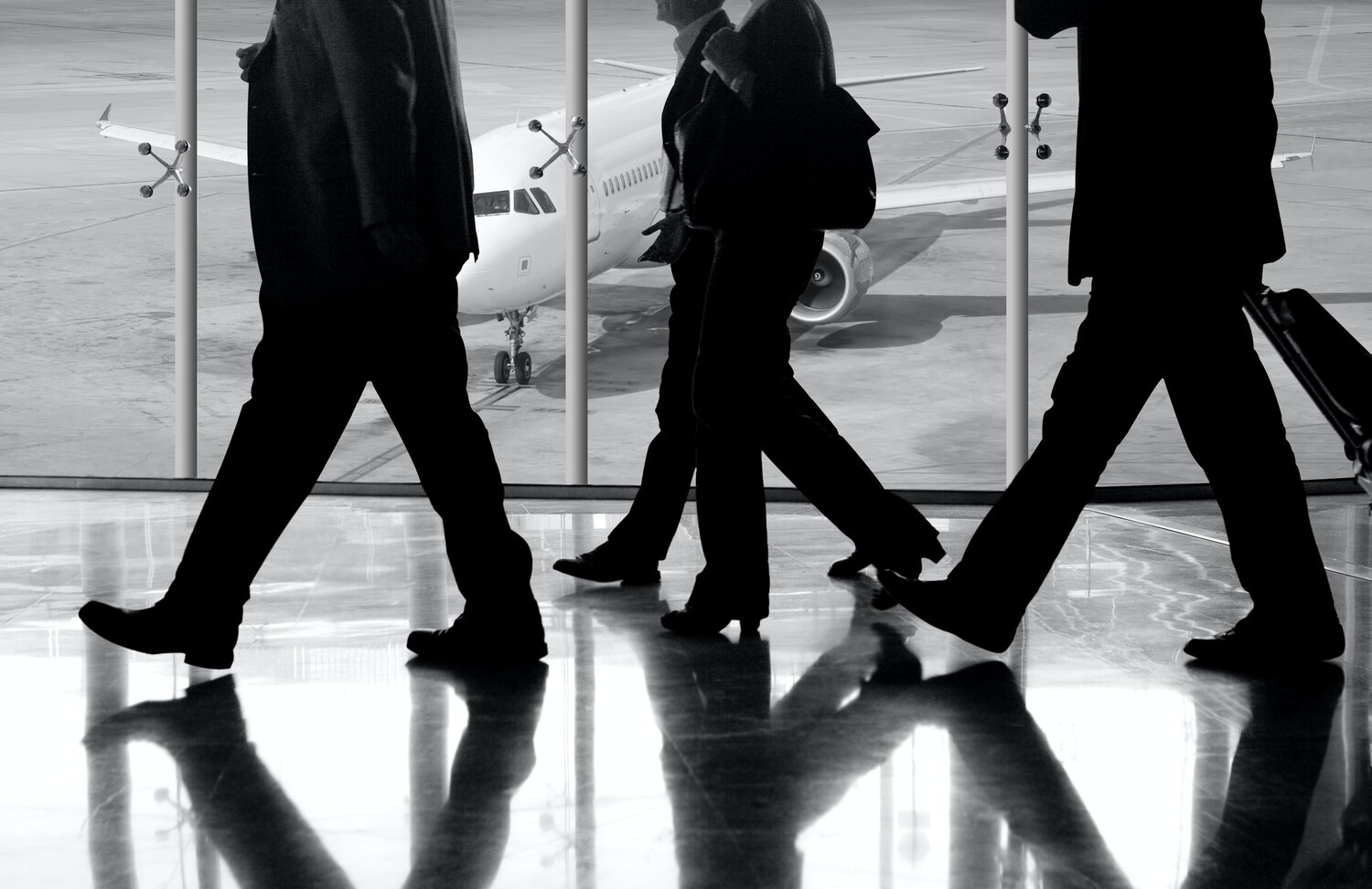 Black and white photo of men in business suits walking in an airport with a plane in the background.