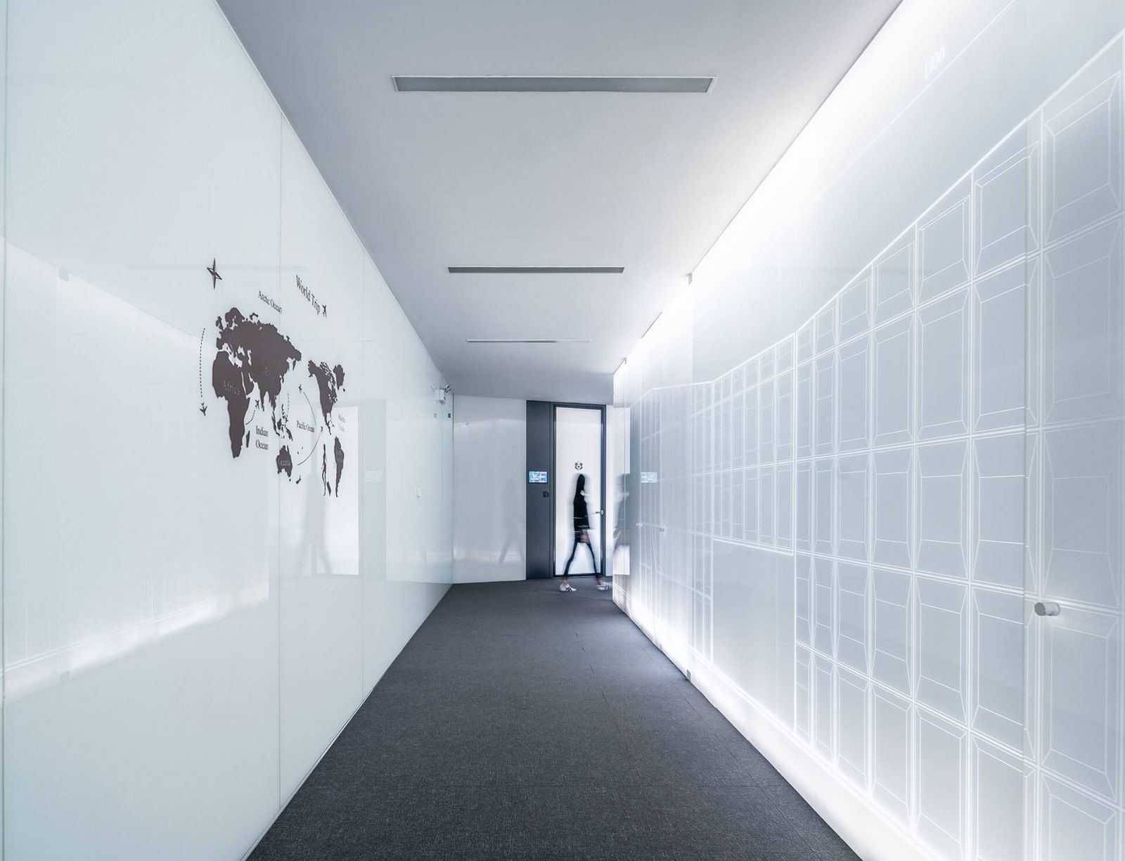 White walled hallway with a world map on the left wall. Woman in background walking around corner traveling to her destination.
